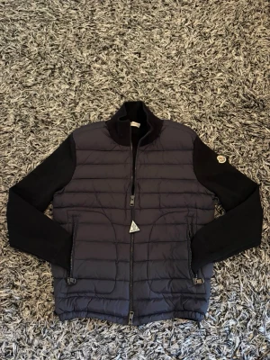 Blå cardigan från Moncler - Snygg blå hybridjacka från Moncler med quiltad front och stickade ärmar. Jackan har hög krage, tvåvägsdragkedja och Moncler-logga på ärmen. Perfekt för kalla vinterdagar och ger en stilren look. Materialmix av dun och stickat tyg för extra komfort.