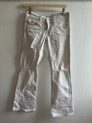 Vita bootcut jeans med slitningar - Säljer ett par vita bootcut jeans med slitningar på knäna och detaljerade fickor både fram och bak. Jeansen har låg midja och klassisk femficksdesign. Perfekta för en avslappnad och trendig look.