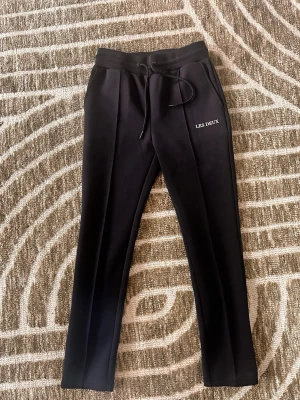 Svarta mjukisbyxor från Les Deux - Snygga mörk blåa mjukisbyxor från Les Deux med raka ben och dragsko i midjan. Byxorna har diskret logga på ena benet och pressveck framtill för en clean look. Tillverkade i ett mjukt och stretchigt material som ger skön passform.