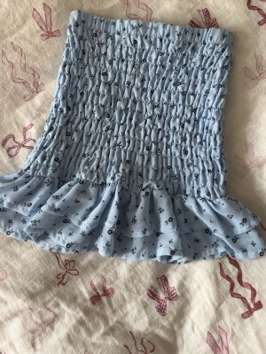 Ljusblå blommig volang tubtopp - Supersöt ljusblå tubtopp med små blommor och volangkant. Toppen är smockad för en tight passform och har en romantisk vibe. Perfekt till sommaren och enkel att styla med jeans eller kjol.