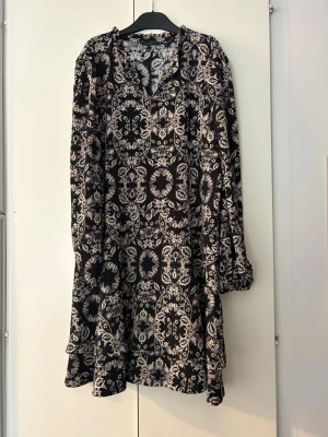 Svartvit mönstrad långärmad klänning strl.L - Svart klänning med vitt blommigt och ornamenterat mönster. Långärmad modell med lös passform och v-ringad hals. Perfekt för dig som gillar bohemiska vibes och vill ha något bekvämt men ändå snyggt. Från Ellos, storlek L.