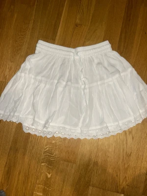 Vit volangkjol med spetskant - Supersöt vit kjol från 157 med volanger och elastisk midja med dragsko. Kjolen har en fin spetskant längst ner som ger en romantisk vibe. Tillverkad i ett lätt och luftigt tyg, perfekt för sommardagar.