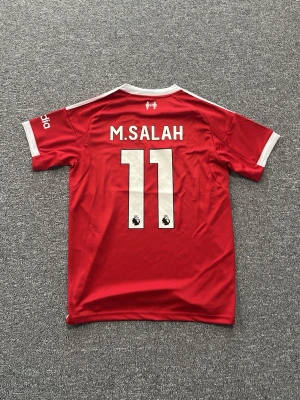 Liverpool matchtröja M.Salah #11 - Röd Liverpool matchtröja med vita detaljer, Adidas-logga och Standard Chartered som sponsor. På ryggen står det M.SALAH och nummer 11. Tröjan har korta ärmar, klubbmärke på bröstet och är tillverkad i ett lätt funktionsmaterial.