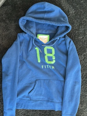 Blå hoodie från Abercrombie & Fitch - Snygg blå hoodie från Abercrombie & Fitch med stor grön broderad 18 och texten 'FITCH' på bröstet. Klassisk känguruficka framtill och justerbar huva med snören. Perfekt för en avslappnad streetwear-look.