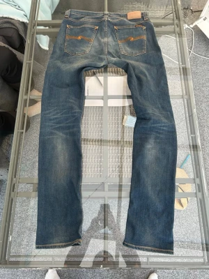 Nudie Jeans Tube Tom W32/L34 – Slim Fit – Turner Shade Blue - Säljer ett par Nudie Jeans Tube Tom i tvätten Turner Shade Blue, storlek W32/L34. Klassiska slim fit-jeans i mörkblå premiumdenim med lätt stretch (98% ekologisk bomull, 2% elastan) för en bekväm passform. Jeansen är i mycket bra skick med endast normalt slitage. Perfekta för dig som söker stilrena herrjeans, designerjeans eller kvalitetsdenim för både vardag och finare tillfällen.