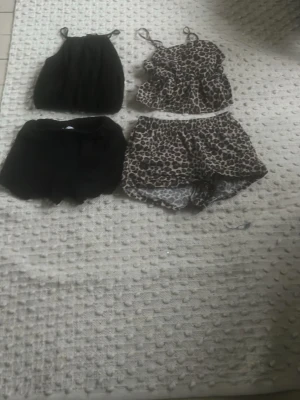2-pack pyjamas-set från Primark - Två pyjamas-set från Primark. Ena setet är helsvart med linne och shorts, det andra har leopardmönster med volanglinne och matchande shorts. Båda seten har tunna axelband och är perfekta för varma nätter. Materialet känns lätt och mjukt mot huden.