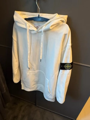 Vit hoodie från Stone Island - Säljer en clean vit hoodie från Stone Island med klassisk patch på vänster ärm. Hoodien har stor huva med dragsko och en rymlig magficka. Tillverkad i mjukt bomullsmaterial som känns skönt mot huden. Perfekt för dig som gillar streetwear och enkel stil.