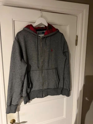Grå zip hoodie från Polo Ralph Lauren - Snygg grå hoodie med dragkedja från Polo Ralph Lauren. Har röd logga broderad på bröstet och röd innerfoder i huvan. Utrustad med kängurufickor och ribbade muddar. Perfekt för en avslappnad och stilren look.