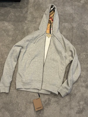 Grå hoodie från Burberry med dragkedja - Snygg grå hoodie från Burberry med dragkedja och klassiskt rutigt foder i huvan. Tillverkad i mjukt bomullsmaterial och har två fickor framtill. Perfekt för dig som vill ha en stilren och exklusiv look med en twist av Burberrys ikoniska mönster.