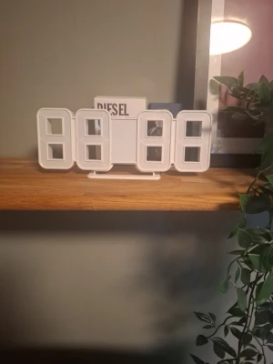 Vit digital LED-väggklocka - Modern digital väggklocka i vit plast med stora LED-siffror som lyser starkt. Klockan har en stilren och minimalistisk design och passar perfekt på hyllan eller väggen. Drivs med el och visar tiden tydligt även i mörker.