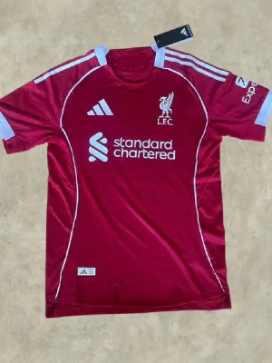 Liverpool matchtröja Salah 11 Adidas - Snygg röd Liverpool matchtröja från Adidas med tryck av M. Salah och nummer 11 på ryggen. Tröjan har vita detaljer, klubbmärke och sponsorlogga framtill samt korta ärmar. Perfekt för dig som älskar fotboll och vill stötta ditt lag.
