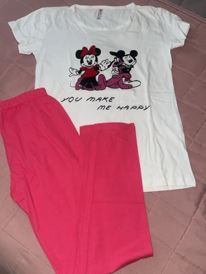 Disney pyjamas med Mimmi & Musse. Oanvänd - Söt pyjamas med vit t-shirt och tryck av Mimmi och Musse Pigg samt texten 'You make me happy'. Tillhörande byxor i starkt rosa färg med resår i midjan. Perfekt för mysiga kvällar och Disneyfans.