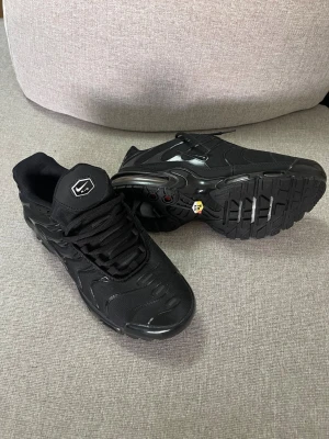 Nike Air Max TN Plus svarta sneakers 43 - Säljer ett par helsvarta Nike Air Max Plus sneakers med mesh och syntetmaterial. Skorna har en chunky sula med synlig Air-enhet och vågiga detaljer på ovandelen. Klassisk Nike-logga på plösen och robust snörning. Perfekta för dig som gillar streetwear.