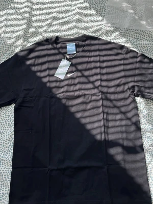 Svart NOCTA t-shirt från Nike - Snygg svart t-shirt från Nike NOCTA-kollektionen med vit swoosh-logga på bröstet och stor NOCTA-tryck på ryggen. Klassisk rund hals och korta ärmar. Tillverkad i mjuk bomull för skön passform. Perfekt för dig som gillar streetwear och stilrena plagg.