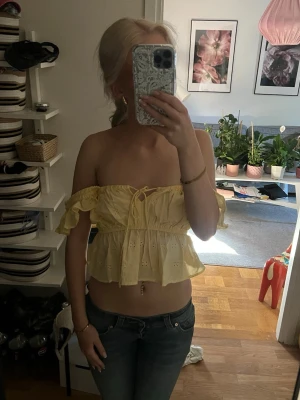 Gul offshoulder volang topp från london  - Söt gul offshoulder topp med volangdetaljer och broderade hålmönster. Toppen har snörning framtill och kan bäras både med axelband eller axelbandslös. Perfekt för sommarens alla tillfällen!