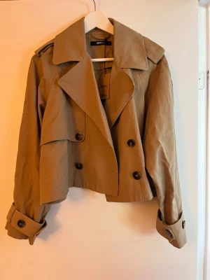 Kort beige trenchcoat från Gina Tricot - Snygg kort trenchcoat i beige från Gina Tricot men köpt av mig på Sellpy och aldrig använd! Jackan är i nyskick med inga defekter alls🥰
