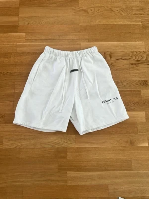 Vita Essentials shorts med snörning - Vita shorts från Essentials med elastisk midja och snörning framtill. Shortsen har fickor på sidorna och en bakficka samt tryckt logga på vänster ben. Tillverkade i mjuk bomull, perfekta för sommaren.