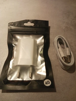 Vit USB-laddare med kabel - Säljer en vit USB-laddare med tillhörande USB-kabel. Laddaren har två metallstift för vägguttag och kommer i en svart och genomskinlig förpackning. Kabeln är vit och har USB-A till USB-C-kontakter. Perfekt för att ladda mobilen eller andra enheter.