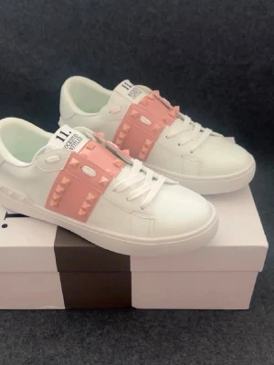 Vita sneakers med rosa nitar - Snygga vita sneakers från Valentino med breda rosa band över vristen dekorerade med pyramidformade nitar. Skorna har klassisk låg profil, vita snören och en chunky sula. Perfekta för dig som vill sticka ut med en cool och trendig detalj.