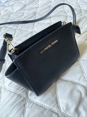 Svart axelväska från Michael Kors - Snygg svart axelväska från Michael Kors i stilren design med guldfärgade detaljer och justerbar axelrem. Väskan är gjord i tåligt läder med struktur och har ett diskret logotyptryck framtill. Perfekt storlek för det viktigaste och enkel att matcha till olika outfits.