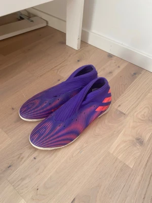 Lila Adidas Nemeziz fotbollsskor - Säljer ett par Adidas Nemeziz fotbollsskor i lila med röda detaljer och vita sulor. Skorna har en strumpliknande design utan snörning och är tillverkade i syntetmaterial för bra passform och kontroll på planen. Perfekta för dig som vill sticka ut på fotbollsplanen!