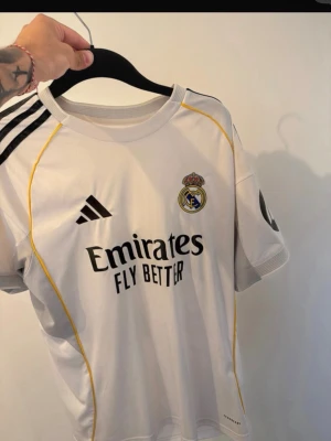 Real Madrid matchtröja Adidas vit - Säljer en vit Real Madrid matchtröja från Adidas med svarta detaljer och gula sömmar. Tröjan har klubbmärke, Adidas-logga och Emirates Fly Better-tryck på bröstet samt svart märke på ärmen. Perfekt för fotbollsfans!