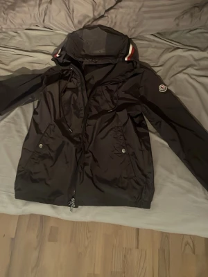 Svart vindjacka från Moncler  - Snygg grå vindjacka från Moncler med huva och dragkedja framtill. Jackan har två fickor med tryckknappar och Moncler-logga på ärmen. Detaljer i rött och vitt på huvan ger en sportig touch. Tillverkad i lätt, vattenavvisande material.