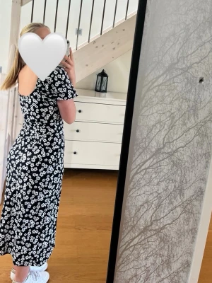 Svart blommig långklänning - Mörkblå långklänning med vita blommor och puffärmar. Klänningen har en fyrkantig ringning, slits framtill och figurnära passform. Perfekt klänning både nu i sommar, men passar även senare under hösten. Klänningen är bara använd ett fåtal gånger, under bröllop och konfirmation.