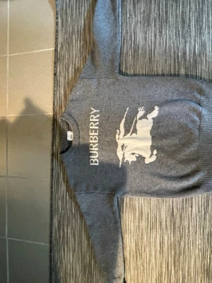 Burberry tröja - Snygg grå stickad tröja från Burberry med stort vitt logotryck och riddarmotiv på bröstet. Tröjan har rund halsringning och ribbade muddar vid ärmslut och nederkant. Perfekt för dig som gillar klassisk design med en modern twist.