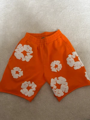 Orange shorts från Denim Tears - Säljer ett par orange shorts från Denim Tears med vita blommönster. Shortsen har elastisk midja och avslappnad passform, perfekta för sommaren. Materialet är mjuk bomull som känns skönt mot huden. Riktigt snygga och unika shorts för dig som vill sticka ut.