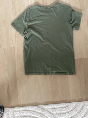 Olivgrön t-shirt från H&M - En enkel olivgrön t-shirt från H&M med rund hals och diskret tryck på bröstet. Klassisk passform och korta ärmar, perfekt till jeans eller shorts. Tillverkad i mjuk bomull för skön känsla hela dagen.