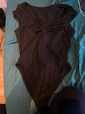 Svart body med kort ärm - En enkel och stilren svart body med korta ärmar och v-spetsad halsringning. Tillverkad i stretchigt material som sitter tajt mot kroppen och har en lätt rynkning vid bysten för extra detalj. Perfekt att matcha med jeans eller kjol.