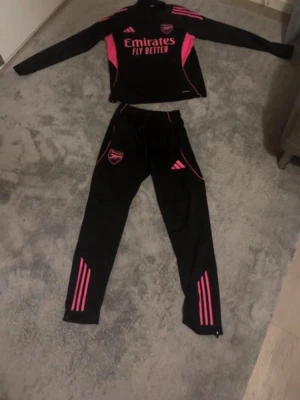 Arsenal träningsset svart/rosa Adidas - Säljer ett Arsenal träningsset från Adidas i svart med rosa detaljer. Setet består av en långärmad tröja och matchande byxor, båda med klubbmärke och Adidas-logga. Materialet är troligtvis polyester, perfekt för träning eller uppvärmning. Snygg design med rosa ränder och tryck.