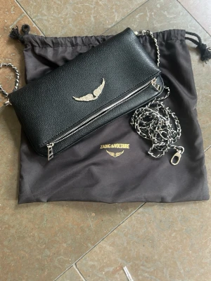 Svart axelväska Zadig & Voltaire - Snygg svart axelväska i läder från Zadig & Voltaire med silverfärgad kedjerem och dragkedjedetaljer. Väskan har ett silverfärgat vingemblem på framsidan och en praktisk ytterficka med dragkedja på baksidan. Perfekt för dig som gillar stilrena accessoarer.