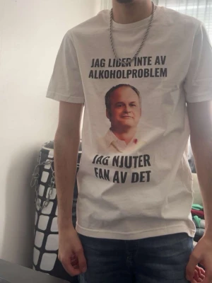 Vit t-shirt med tryck och text - Vit t-shirt med svart text och ett tryck på bröstet. Texten lyder 'Jag lider inte av alkoholproblem, jag njuter fan av det'. Klassisk passform med rund hals och korta ärmar. Perfekt för dig som gillar humoristiska plagg.