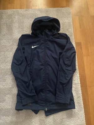 Mörkblå Nike regnjacka med huva - Säljer en mörkblå regnjacka från Nike med huva och hel dragkedja framtill. Jackan har ett vitt Nike-logotyp på bröstet och är tillverkad i ett vattenavvisande material. Perfekt för regniga dagar och sportiga aktiviteter.