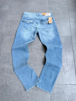 True Religion herr - Säljer ett par ljusblå jeans från True Religion med raka ben och klassiska kontrastsömmar. Jeansen har två bakfickor med lock och knapp samt läderpatch med logga i midjan. Tillverkade i mjuk denim med snygg tvätt.