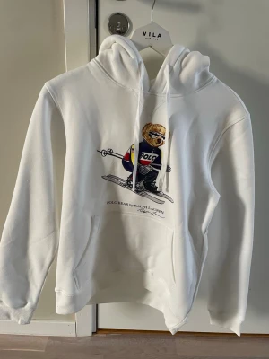 Vit hoodie Polo Bear Ralph Lauren - Säljer en vit hoodie från Ralph Lauren med den ikoniska Polo Bear-trycket framtill där björnen åker skidor. Hoodien har huva med dragsko och en stor magficka. Mjuk och skön bomullsblandning, perfekt för chill dagar. Storlek M kan passa S