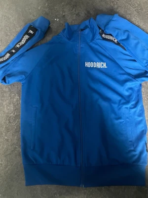 Blå zip hoodie från Hoodrich - Snygg blå hoodie från Hoodrich med dragkedja framtill och vita logotexter på bröstet och längs ärmarna. Hoodien har ribbade muddar och två fickor på sidorna. Tillverkad i mjukt material som känns skönt mot huden. Perfekt för en streetwear-look.