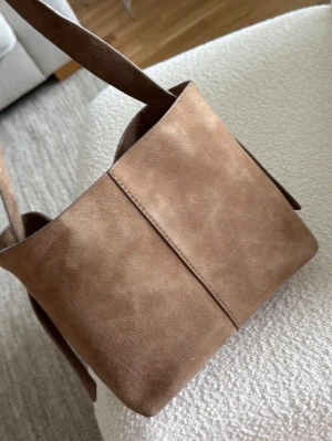 Beck söndergaard fraya - Snygg handväska i mjuk brun mocka med bred axelrem och stilren bucket-modell. Väskan har minimalistisk design med synlig mittsöm och öppet huvudfack. Perfekt för dig som gillar en enkel men trendig look.