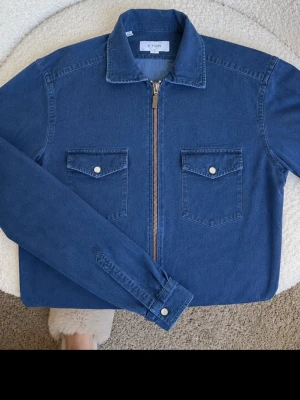    ETON Denim Jacket Slim Fit Strl 38 / S - ETON denim jacket i mörkblå tvätt. Slim fit, storlek 38 / S. Stilren modell med dragkedja framtill och två bröstfickor med tryckknappar. Perfekt som lätt jacka eller overshirt under vår, sommar och tidig höst.  • Märke: ETON • Storlek: 38 / S • Passform: Slim Fit • Färg: Mörkblå denim • Stängning: Dragkedja • Skick: Mycket fint skick, se bilder  Säljs då den inte kommer till användning. Skickas snabbt och väl emballerat.
