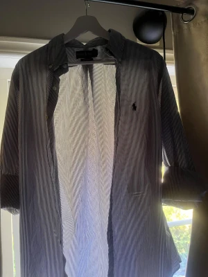 Ralph Lauren skjorta - Säljer en klassisk blåvit randig skjorta från Polo Ralph Lauren med broderad logga på bröstet. Skjortan har lång ärm, knappar framtill och krage. Perfekt för dig som gillar stilrena plagg med ikonisk design.