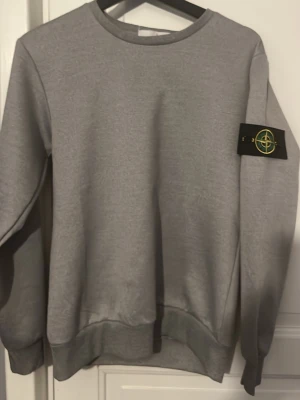 Grå sweatshirt från Stone Island - Snygg grå sweatshirt från Stone Island med klassisk rund hals och ribbade muddar. Tröjan har den ikoniska Stone Island-loggan på vänster ärm och är tillverkad i mjukt bomullsmaterial. Perfekt för en clean och stilren look.