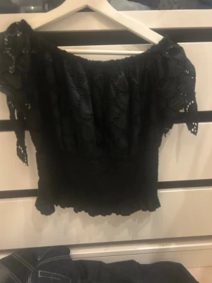 Svart offshoulder blus med spets - Snygg svart offshoulder blus med spetsdetaljer och korta ärmar. Blusen har ett elastiskt liv och volangkant nedtill, perfekt för en trendig look. Luftigt material och fina hålmönster ger en romantisk känsla.