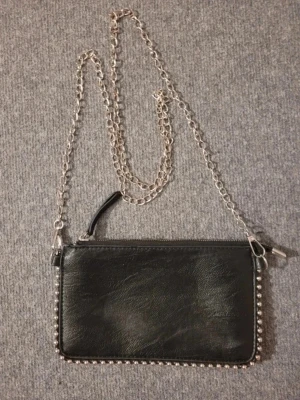 Svart kuvertväska med silverkedja - Snygg svart kuvertväska i fejkläder med dekorativa silverfärgade nitar runt kanterna och en lång silverkedja som axelrem. Väskan har dragkedja upptill och minimalistisk design, perfekt för att lyfta vilken outfit som helst.
