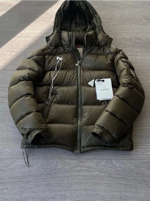 Olivgrön dunjacka från Moncler - Snygg olivgrön dunjacka från Moncler med huva och dragkedja framtill. Jackan har puffig design, två sidofickor med dragkedja och Moncler-logga på ärmen. Tillverkad i glansigt material och fylld med dun för extra värme. Perfekt för kalla vinterdagar.