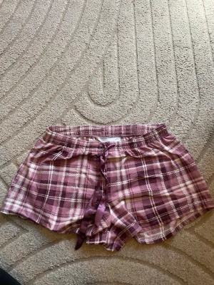 Rutiga pyjamasshorts från Isabella - Mysiga pyjamasshorts från Isabella i lila och vitt rutigt mönster. Shortsen har en bekväm resår i midjan och ett lila satinband för justering. Perfekta för chillkvällar hemma.