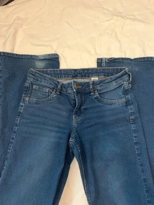 Blå bootcut jeans med stretch - Säljer ett par klassiska blå bootcut jeans med fem fickor och dragkedja. Jeansen har en snygg tvätt och är tillverkade i stretchigt denimtyg för extra komfort. Dem har tecken på användning längst ned benen men inget tyg som är grovt skadat