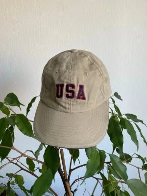 Vintage Keps - Snygg beige keps med böjd skärm och broderad text 'USA' i rött och blått framtill. Klassisk sexpanel-design med ventilationshål och justerbar passform bak. Tillverkad i bomullstyg som ger en avslappnad och sportig look.