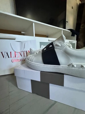 Valentino Garavani vita sneakers - Snygga vita sneakers från Valentino Garavani med svart detalj på sidan och nitar på hälen. Skorna har en klassisk låg siluett, vita snören och en robust gummisula. Perfekta för dig som vill ha en stilren och exklusiv look.